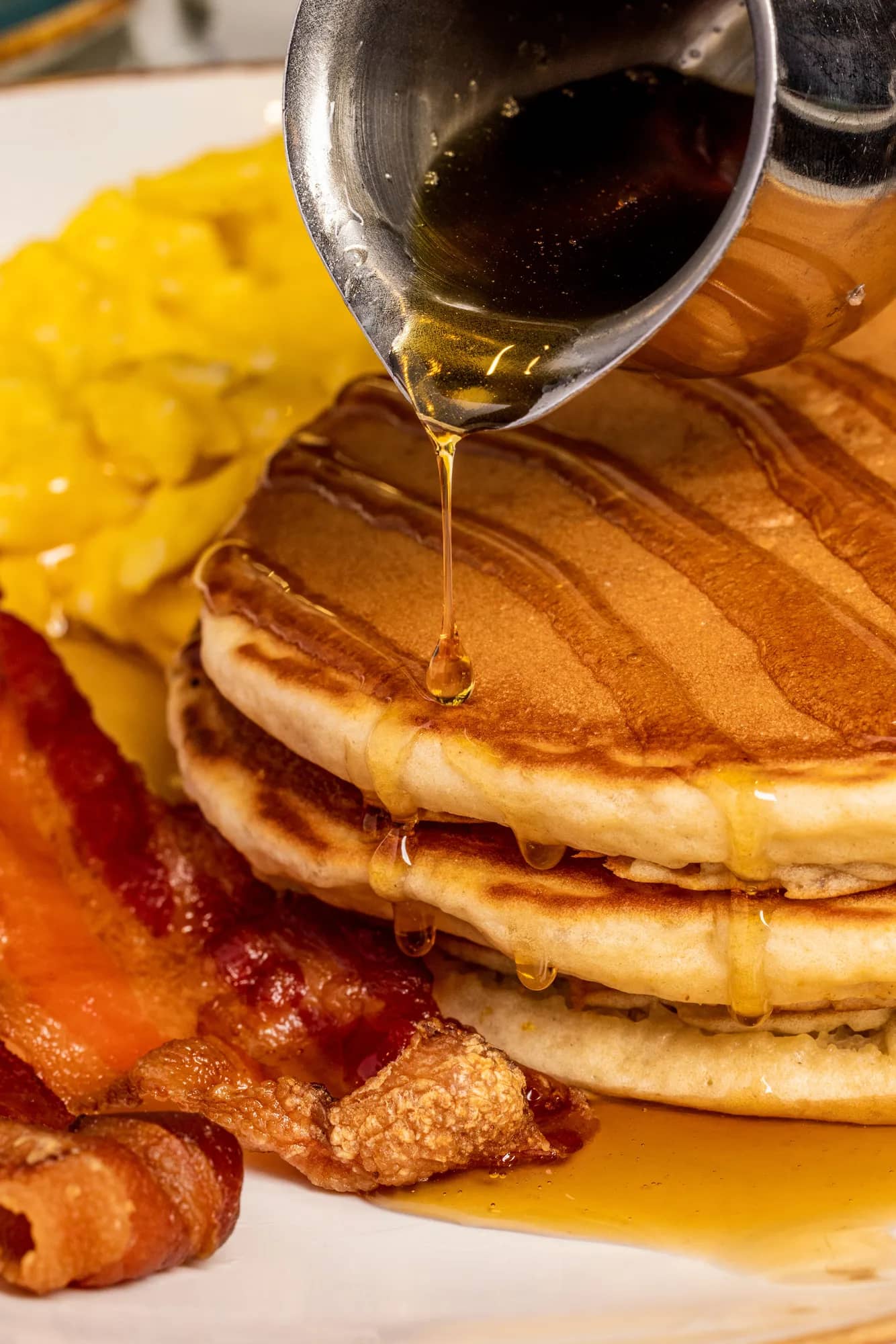 Maple syrup drizzling over a stack of pancakes with scrambled eggs and bacon
